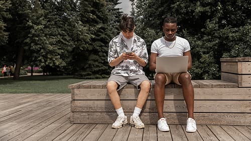 Two men using mobile devices and laptop sitting on a bench