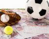 baseball glove and ball, soccer, tennis and rugby balls near betting lists on euro and dollar banknotes